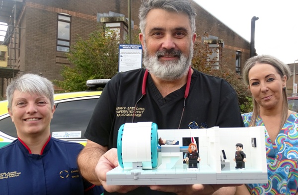 Three people outside a hospital holding a model of an MRI scanner