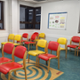 Image shows the back of the children’s waiting room with 8 pairs of red and yellow chairs placed in the room. There are brown framed windows along the side with a nutrition information board on the back wall.