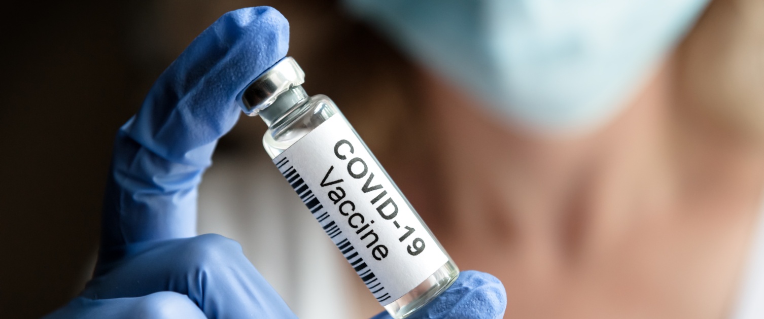 A gloved hand holds a vial marked COVID vaccine.