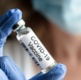 A gloved hand holds a vial marked COVID vaccine.
