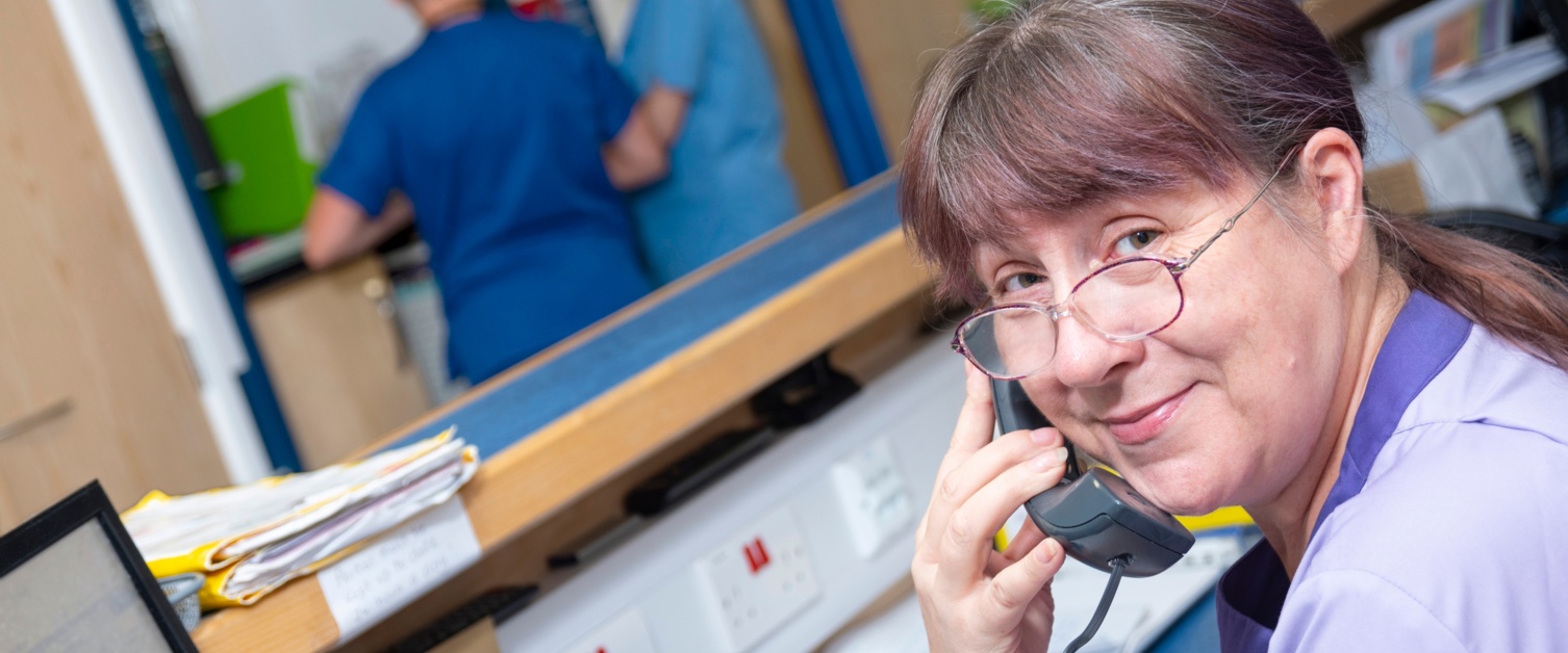 A nurse smiling at the camera whilst on the phone with two nurses in the background.