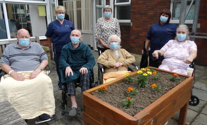 Image shows people standing and sitting behind a flower bed
