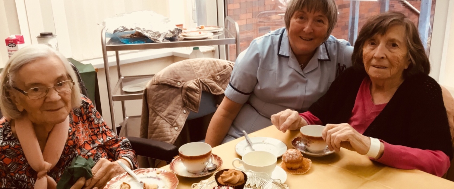 An elderly lady enjoys a cup of tea with her daughter in front of a table covered with a yellow cloth and laid with china cups and saucers.