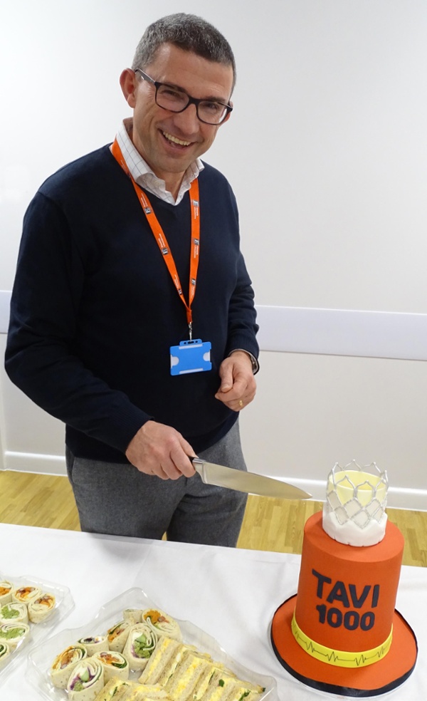 Image shows a smiling man using a knife to cut a celebration cake