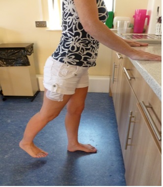 A patient stands facing a kitchen unit with their hands on the unit. Their right leg is extended slightly behind them.