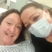 Twins take a selfie. One is lying in a hospital bed with an oxygen cannula under her nose while the other leans in wearing a surgical mask.