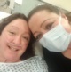 Twins take a selfie. One is lying in a hospital bed with an oxygen cannula under her nose while the other leans in wearing a surgical mask.