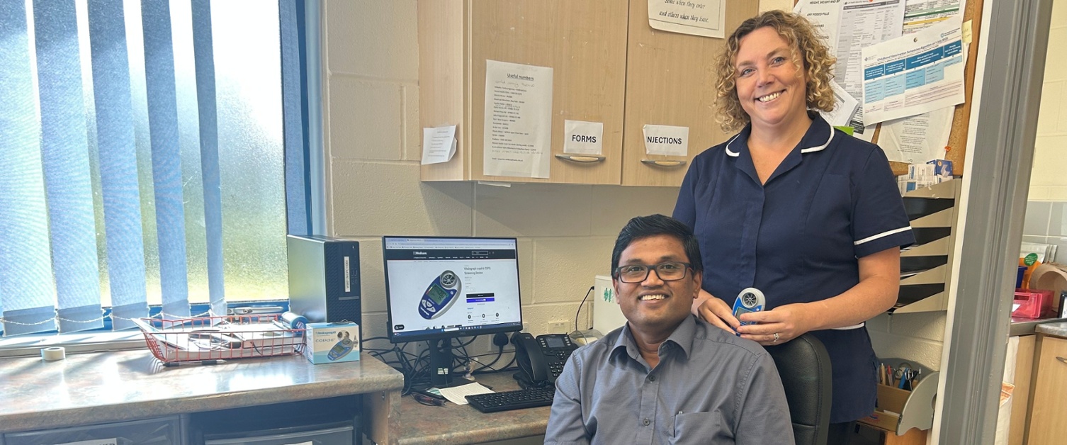 Kannan yn eistedd i lawr a Rebecca yn sefyll y tu ôl iddo yn dal y ddyfais