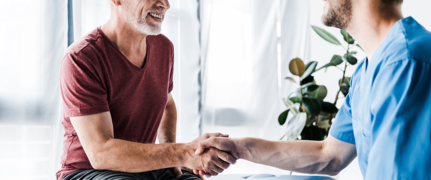 Claf yn eistedd ar ochr wely mewn siorts a chrys yn ysgwyd llaw gyda meddyg yn gwisgo gwisg las. Mae