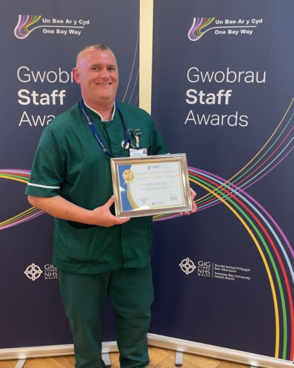 Steve yn gwisgo ei wisg ac yn dal ei dystysgrif wobr