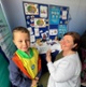 Delwedd o fachgen ifanc a menyw yn gwenu ar y camera, yn sefyll o flaen stondin arddangos nyrsio ysgol.