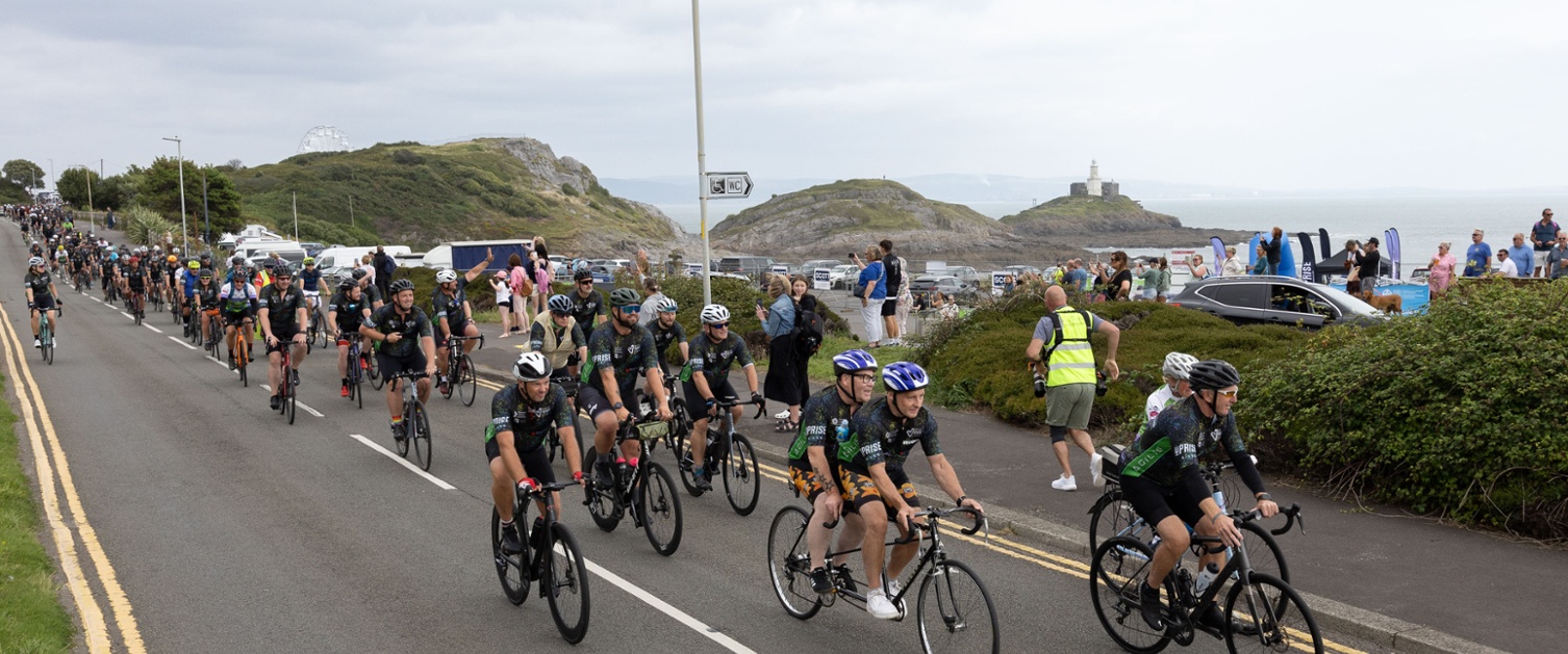 Grŵp mawr o feicwyr yn seiclo heibio goleudy, gyda Jonathan Davies o flaen