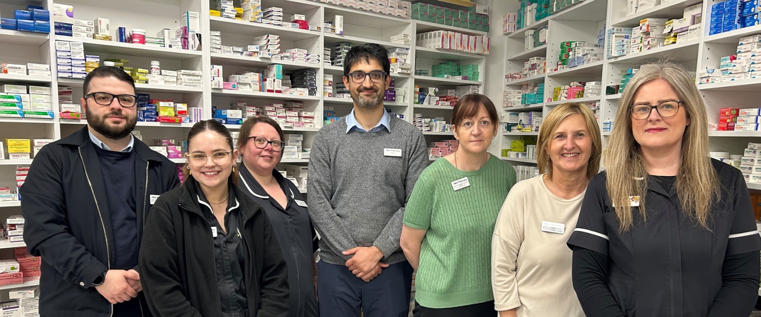 Aelodau staff o Allied Pharmacy yn sefyll o flaen y cwpwrdd meddyginiaethau