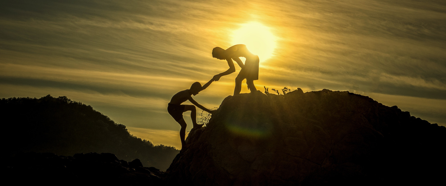 A person helps another climb a hill while the sun sets in the background