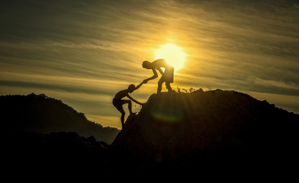 A person helps another climb a hill while the sun sets in the background
