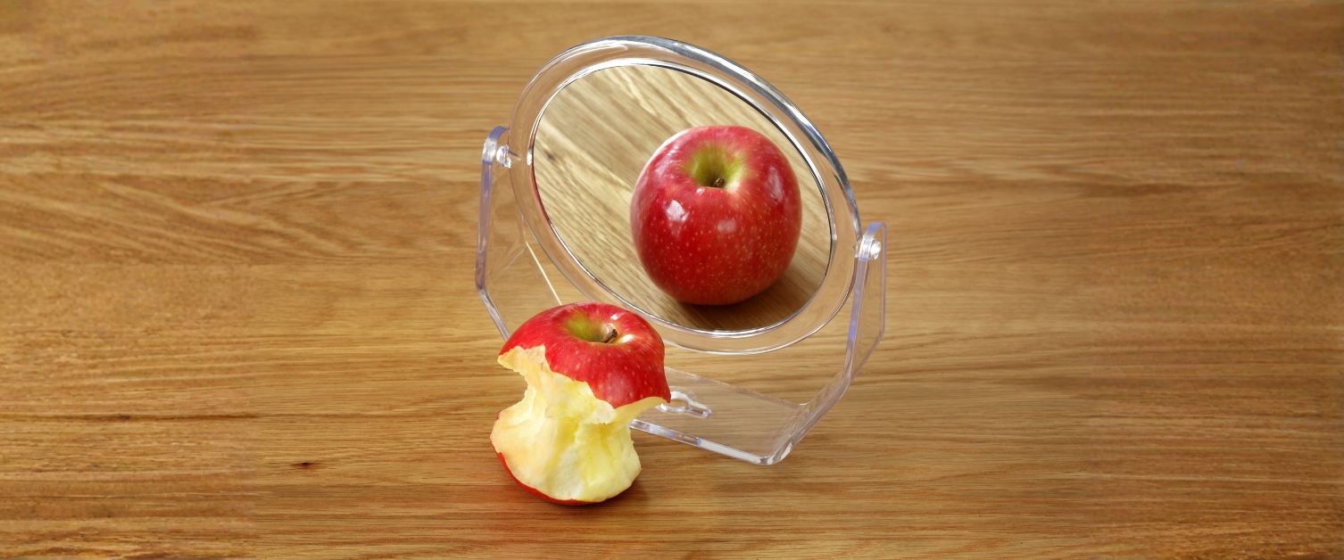 A bitten apple reflected as whole in a mirror on a table
