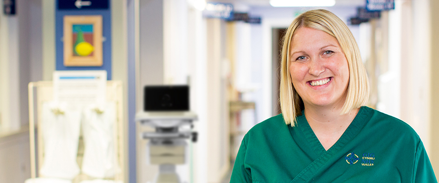 Blonde, smiling nurse in NHS Wales uniform standing in hospital corridor