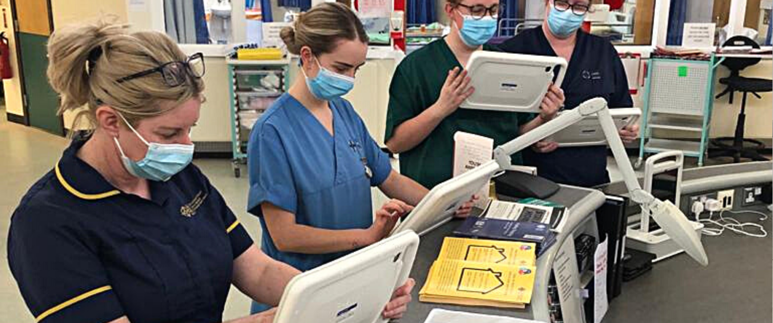 4 nurses stand at a curved desk, each one holds a tablet that displays the Welsh Nursing Care Record. They