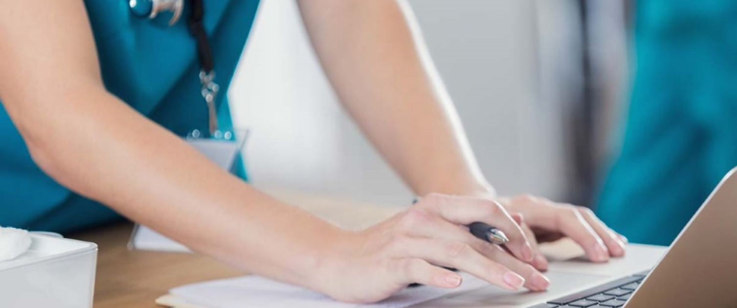 Female clinician wearing blue scrubs leans over a computer.