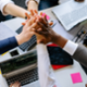 Five people giving each other a group high five over a table, over a laptop and post it notes.