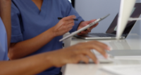 A close up of the torsos and arms of two clinicians using a tablet and a computer. They are wearing blue scrubs.
