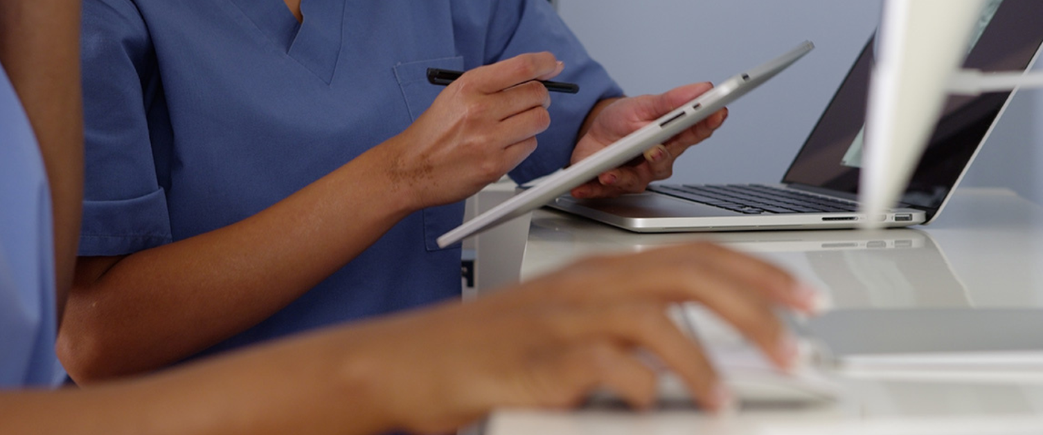 A close up of the torsos and arms of two clinicians using a tablet and a computer. They are wearing blue scrubs.