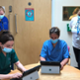 Four nurses, two sitting and two standing, look at laptops that display the Welsh Nursing Care Record while wearing masks