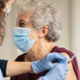Older woman wearing a mask has her sleeve rolled up and is about to receive a vaccination from a clinician wearing blue gloves