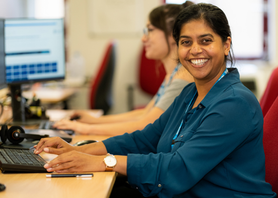 Smiling woman with dark hair looks at the camera as she works at her desk, with a woman with glasses in the background working at her computer