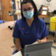 A brown haired nursing wearing glasses and a mask looks at the camera while sitting in front of a laptop that displays the Welsh Nursing Care Record