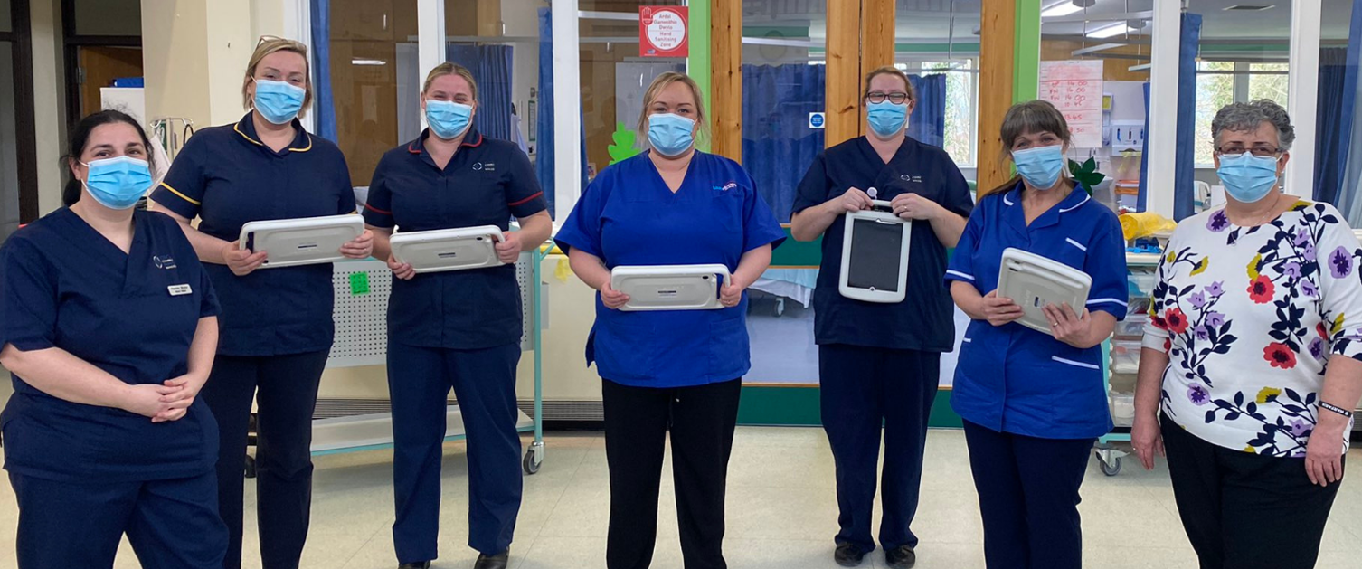 A group of nurses stand together in a hospital wearing masks and holding tablets that feature the Welsh Nursing Care Record