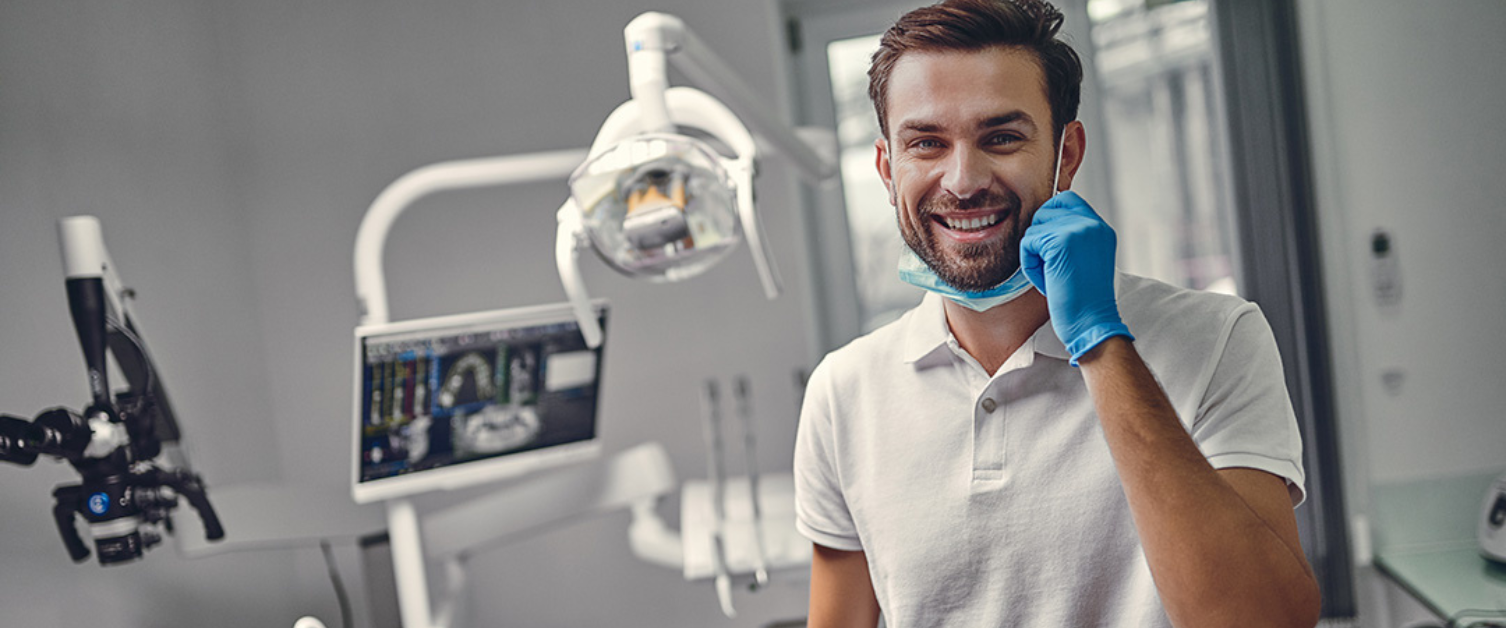 Dentist smiling at the camera wearing gloves and a mask, the background shows that he