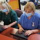 Two nurses, one in green scrubs and one in blue scrubs, sit at laptops using the Welsh Nursing Care Record