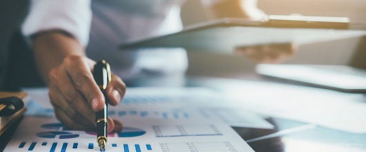 Business person looking over spreadsheets laid out on a desk. They are holding a clipboard and a pen. 