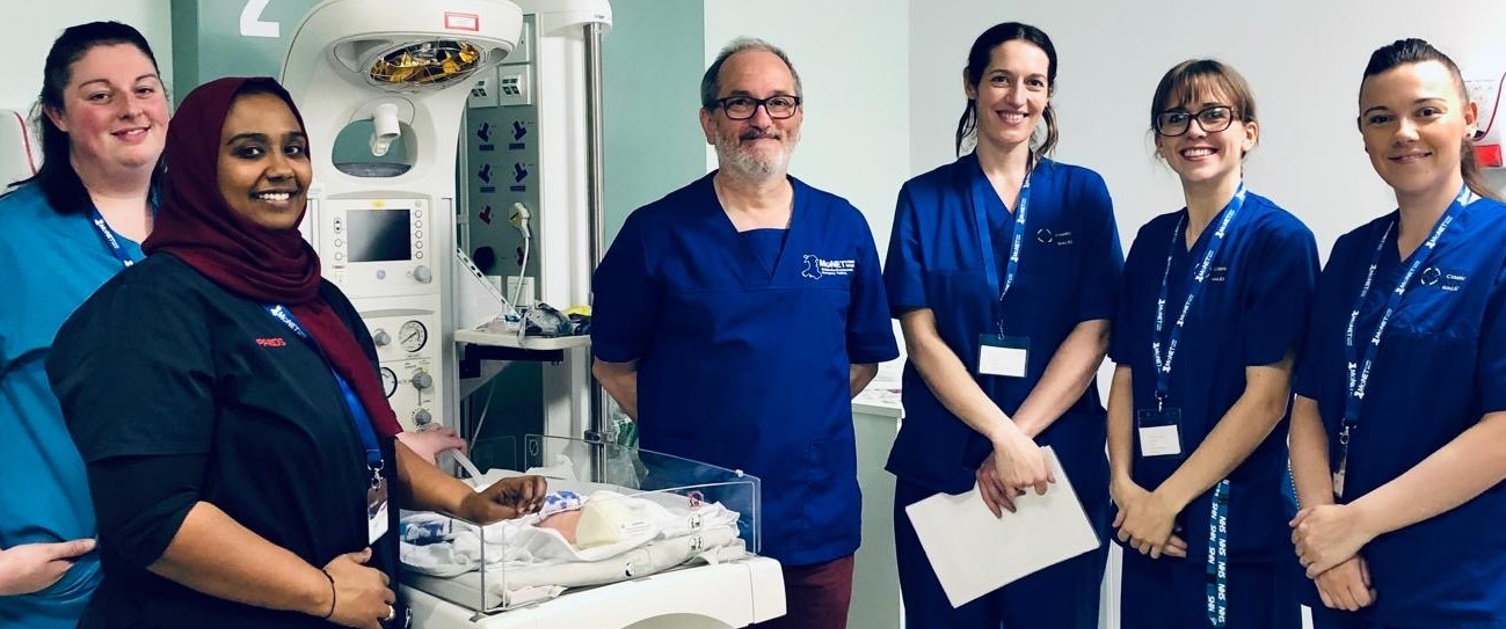 MoNET Wales Dr Iyad Al-Muzaffar in MoNET royal blue scrubs standing with the neonatal team in front of a neonatal resuscitaire.
