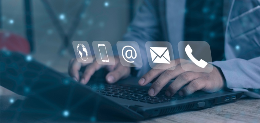 Person typing on a computer keyboard in the background. In the foreground there are the symbols for contacting something. i.e. a telephone, an email, an @ sigh and a world icon.