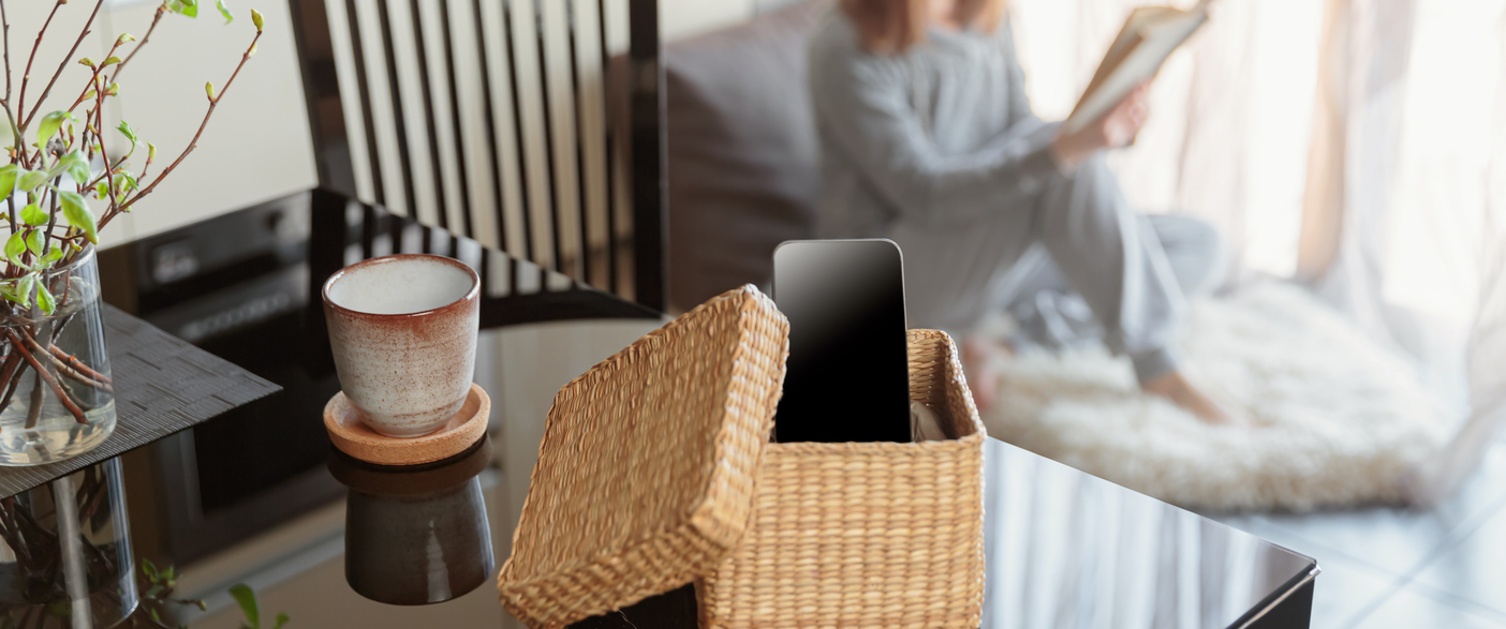 Woman sitting near window and reading book. Focus on box with smartphone. Digital detox and leisure concept