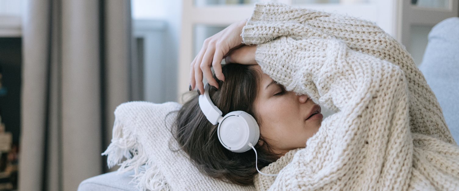 Image of a lady lying down on a sofa wearing a woolly jumper, listening to something on her headphones, with her hands resting on her forehead.