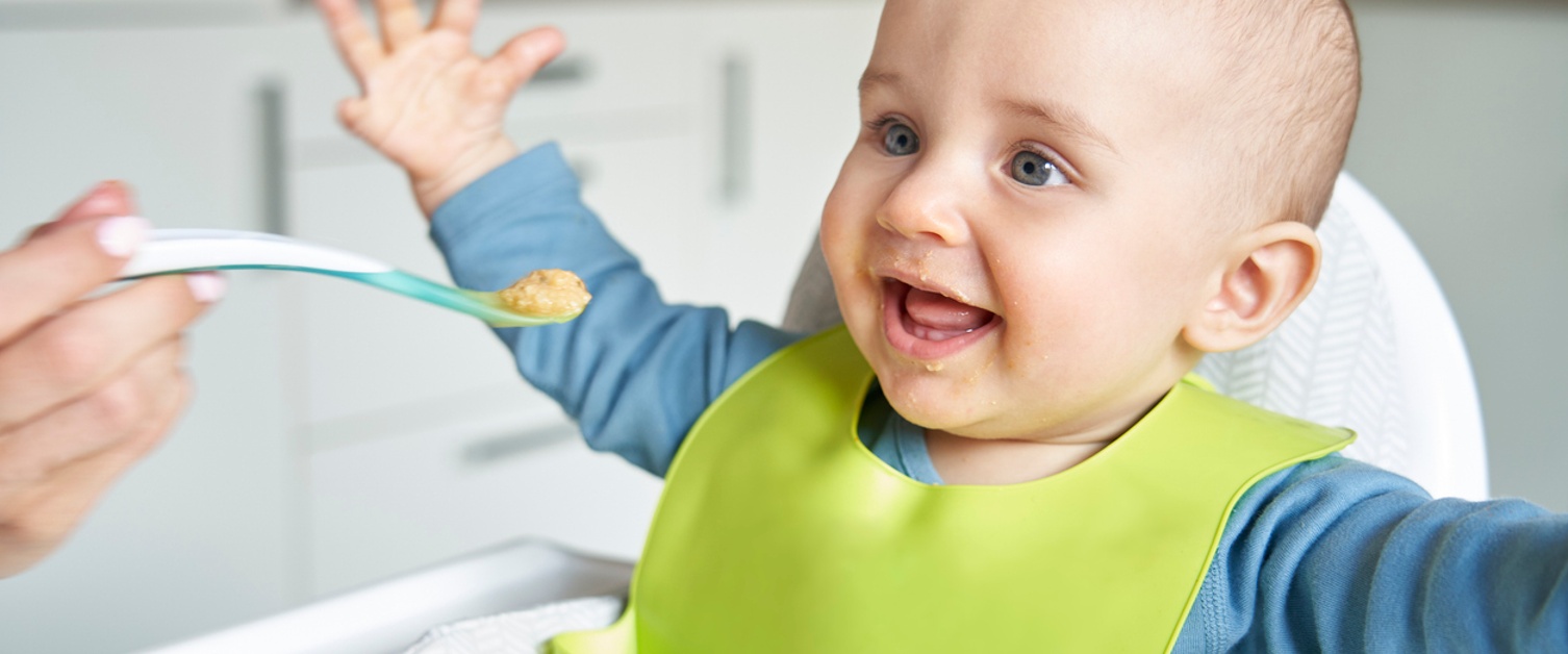 Smiling 8 month Old Baby Boy At Home In High Chair Being Fed Solid Food By Mother With Spoon