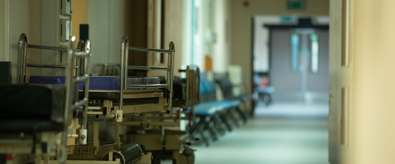 Copilot said:  English Alt Text  Hospital corridor with patient trolleys and seating lined along the hallway, leading towards double doors in the distance.