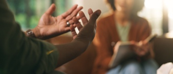 Close-up of people in discussion, with one person gesturing while speaking and another taking notes in the background.