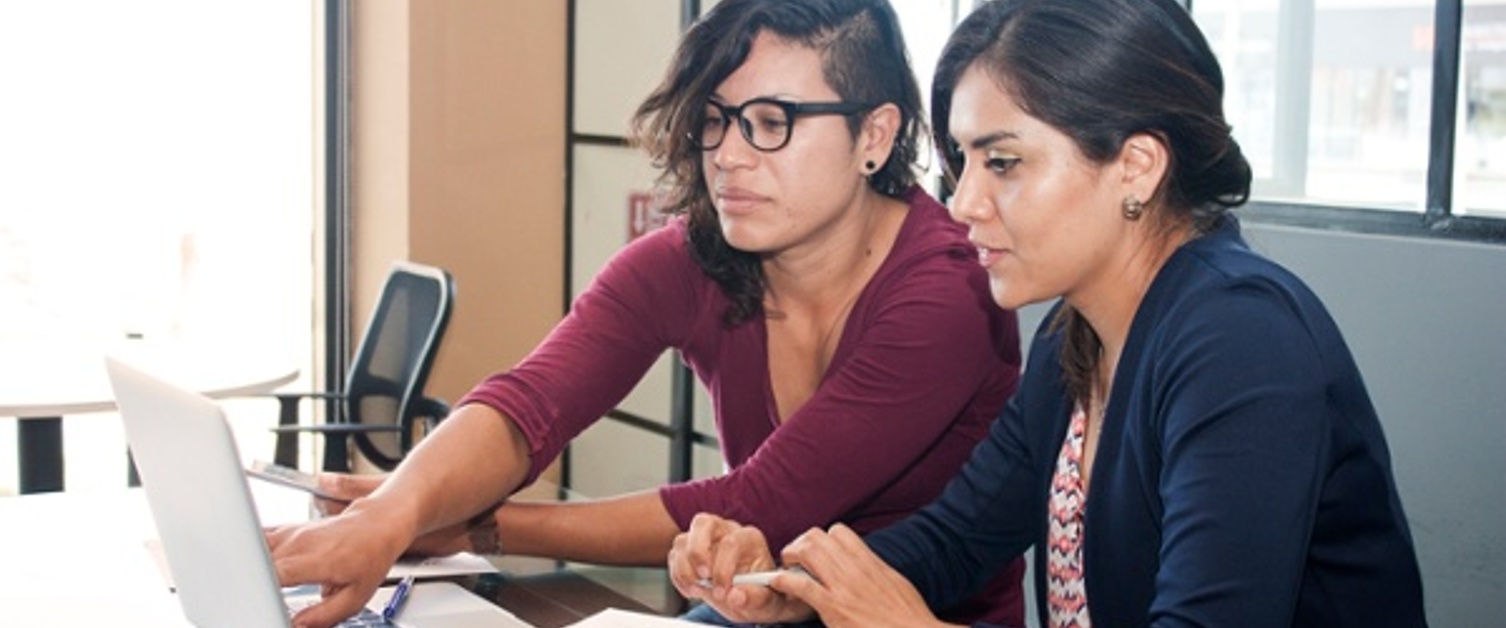 Two young women - one wearing glasses - working together at a table in an office, looking at a silver laptop computer. One is pointing at the computer screen. A pen, a piece of paper and a mobile phone are also on the table.