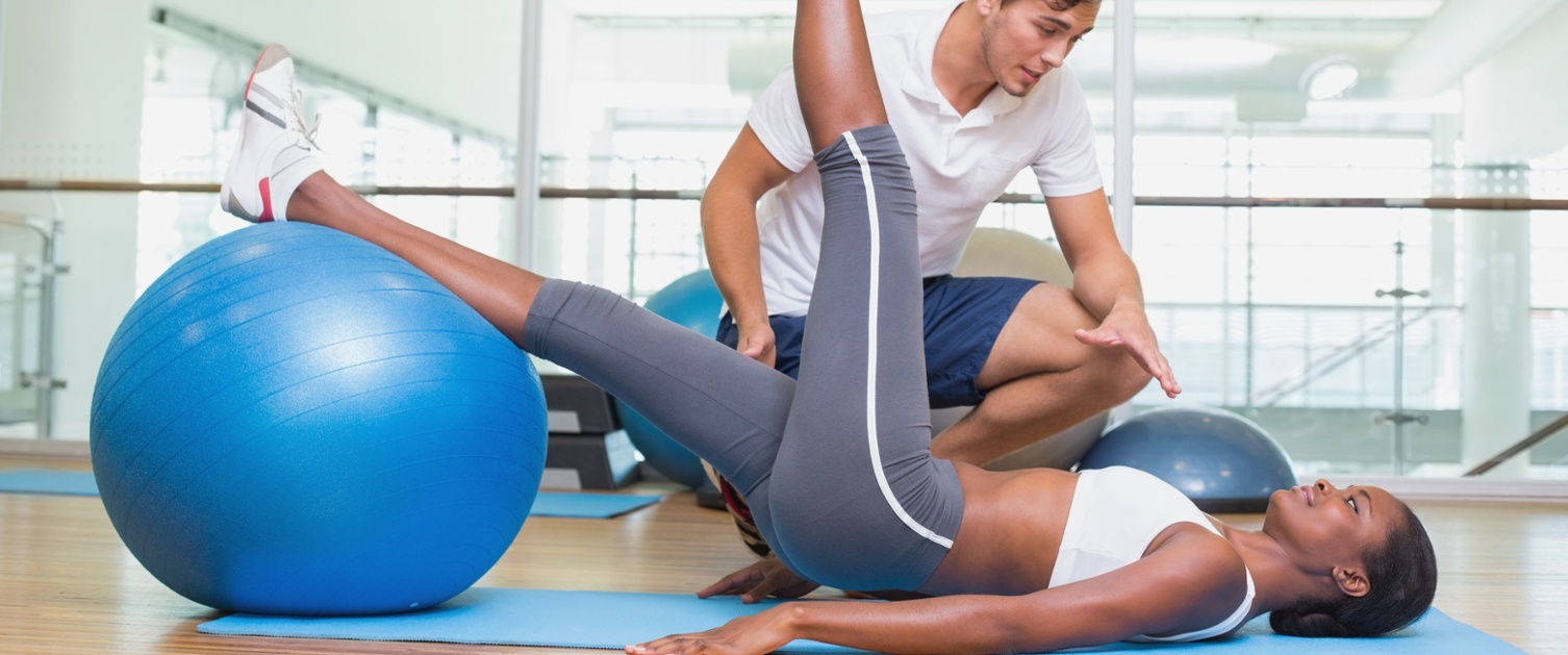 Ffisiotherapydd mewn crys gwyn yn cyflwyno claf ifanc i ymarferion mewn campfa. Mae