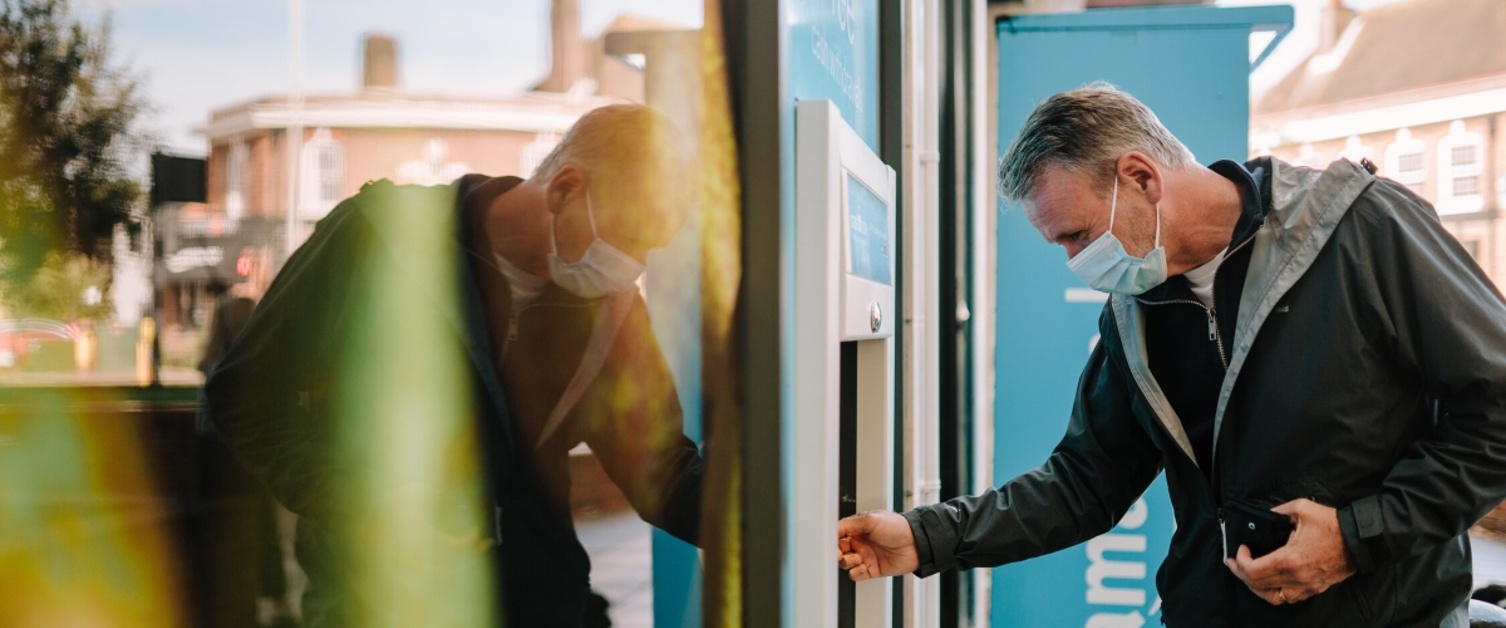 A man is using an ATM cash machine. He is wearing a face mask.