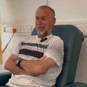 A man is smiling as he is sat in a chair on a hospital ward.