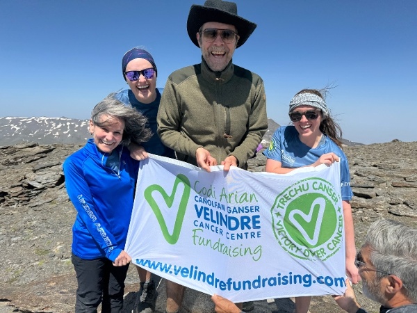 A group of people are holding a Velindre Cancer Charity flag and smiling on top of a mountian.
