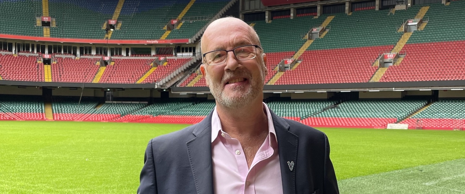 Professor Tom Crosby OBE stands on the pitch at the Principality Stadium. He is wearing a suit, smiling and happy.