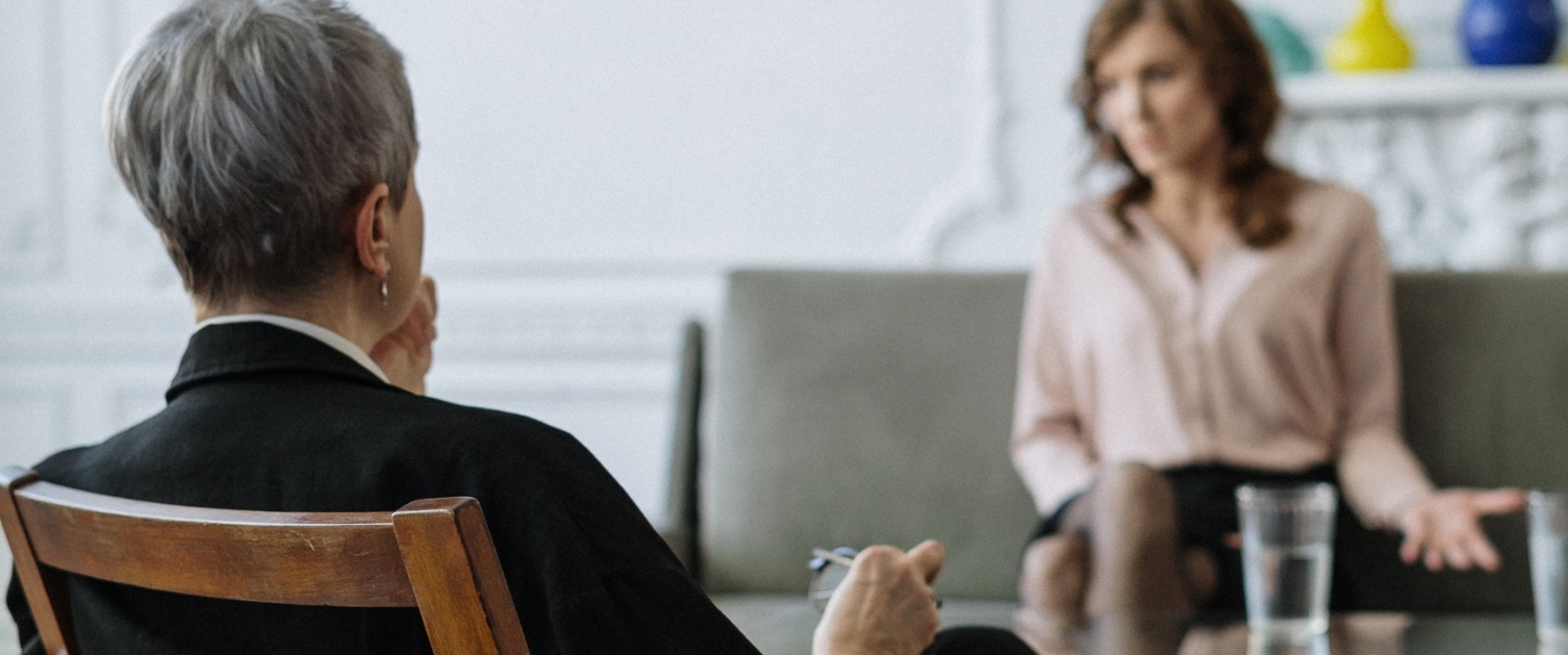 A counsellor is sat on a chair in the foreground, while a woman is sat on a sofa opposite.