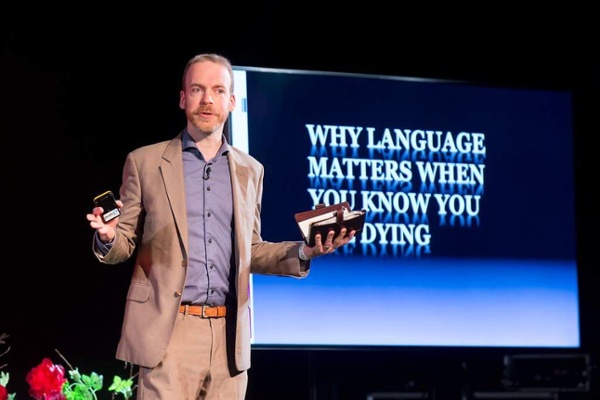 A man is stood on a stage in front of a screen that says: "Why language matters when you know you are dying."
