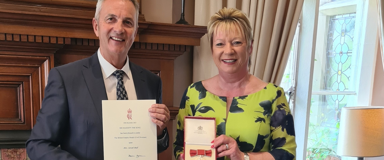 The Lord-Lieutenant and Sarah Bull smile as they hold the British Empire Medal.